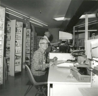 Unidentified man at a desk with medicines on shelves behind him