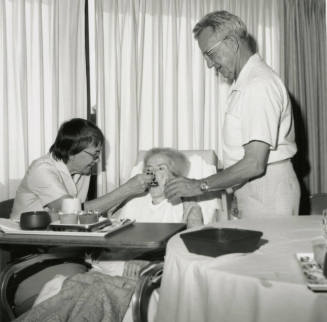 Man and woman feed ill elderly woman in a hospital bed, January 21-27, 1985