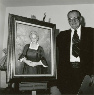 An man holding two paintbrushes stands next to a portrait of an unknown woman