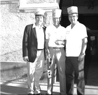 3 men stand outside a concrete building, wearing hats