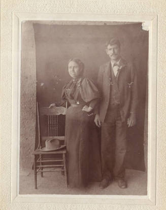 Photograph of a woman and a man standing in dark clothing by a Victorian wooden chair with black cloth pinned to the back wall.