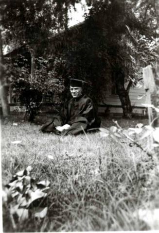 Frank Raymond in his cap and gown, sitting on a lawn in front of a house.