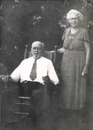 Dr. Fenn J Hart sitting in a rocking chair under a tree, Mrs. Rosa Brown Hart stands to his left