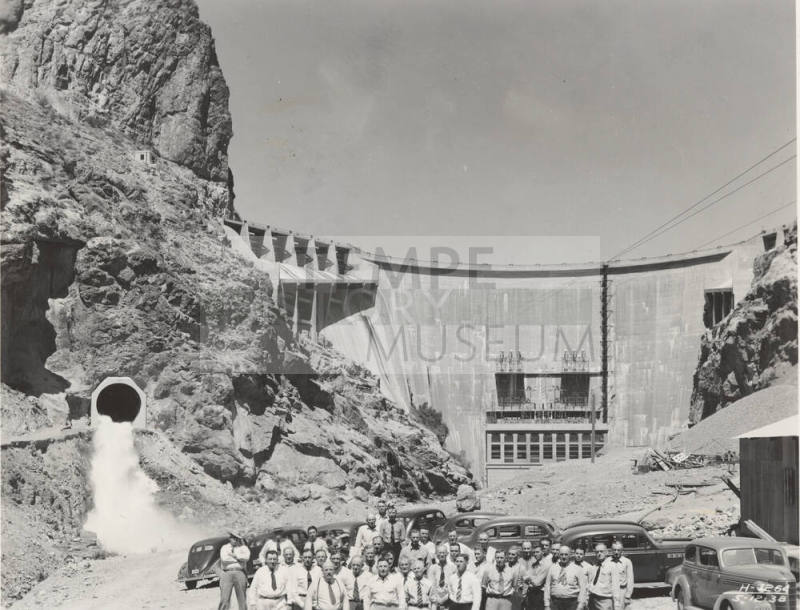 Photo- A row of thirty six men standing in front of Horse Mesa Dam with cars