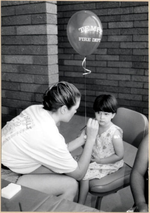 Black and white photograph of Fire Prevention Day of girl with balloon being face painted