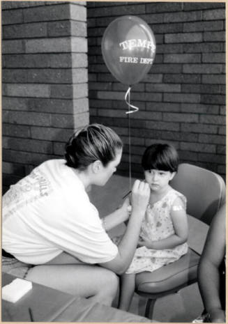 Black and white photograph of Fire Prevention Day of girl with balloon being face painted