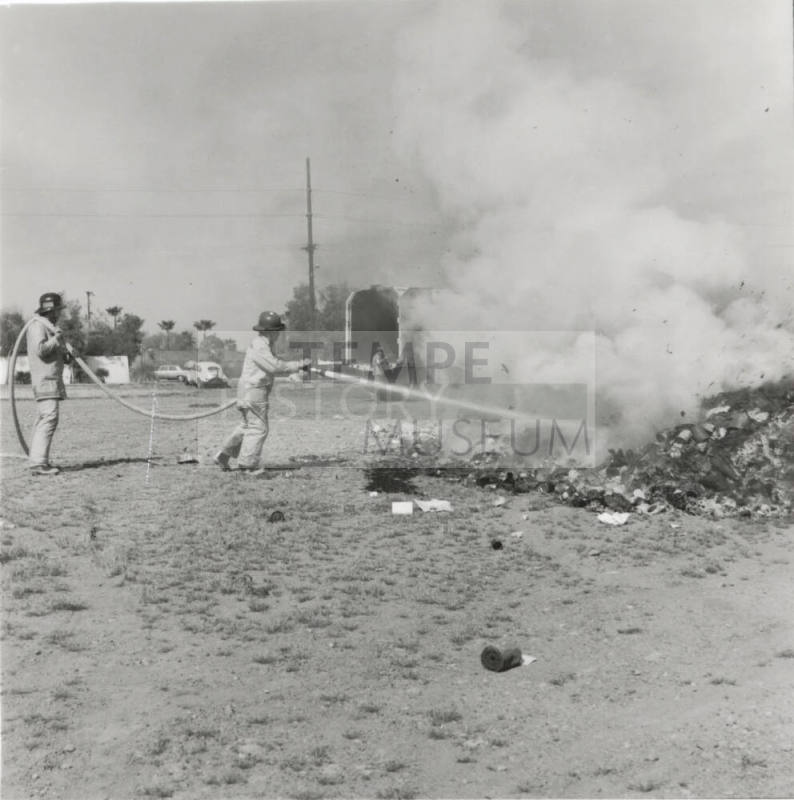 Two Firemen Hose a Smoldering Pile