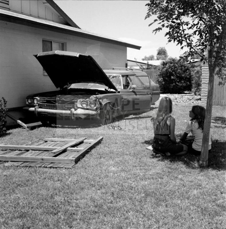 Two people sit under a tree near a wrecked vehicle (photo 1 of 4)-July 1977.
