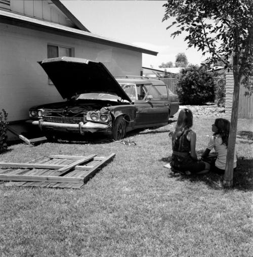 Two people sit under a tree near a wrecked vehicle (photo 1 of 4)-July 1977.