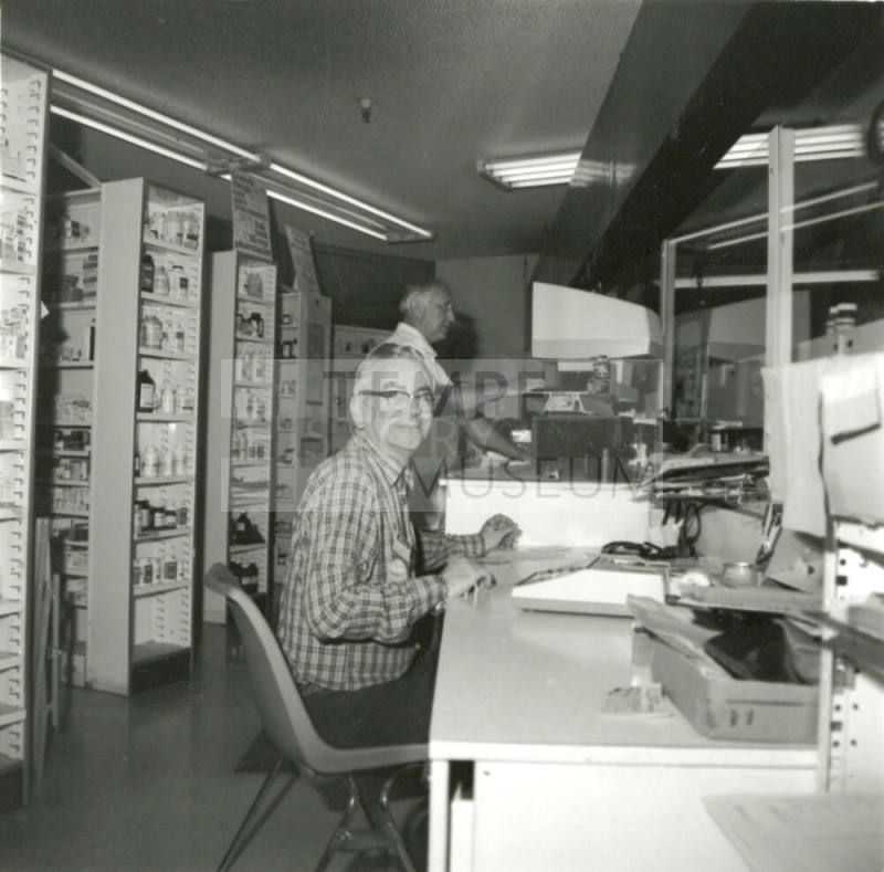 Unidentified man at a desk with medicines on shelves behind him