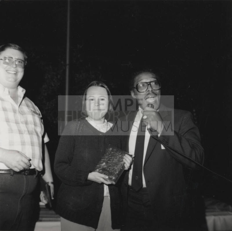 Woman poses with book with a smiling man and a man with a microphone