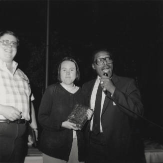 Woman poses with book with a smiling man and a man with a microphone
