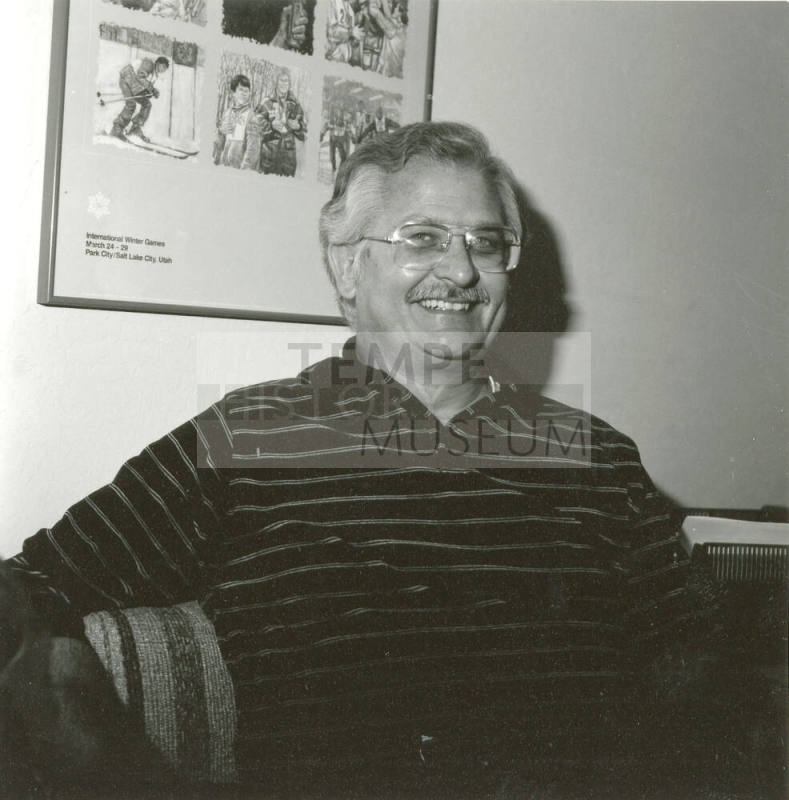 Seated man smiling in front of International Winter Games poster