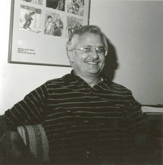 Seated man smiling in front of International Winter Games poster