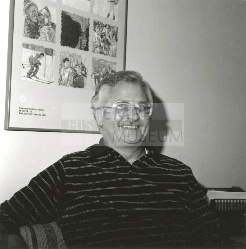 Seated man smiling in front of International Winter Games poster