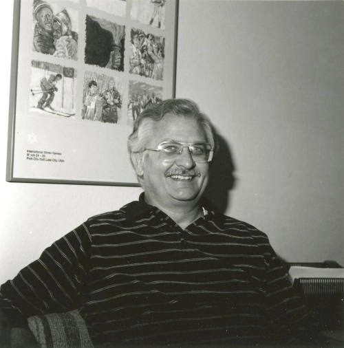 Seated man smiling in front of International Winter Games poster