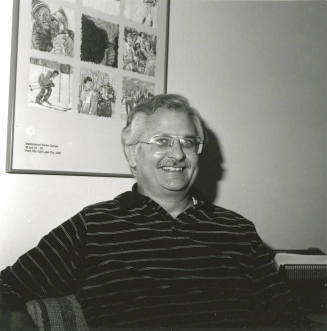 Seated man smiling in front of International Winter Games poster