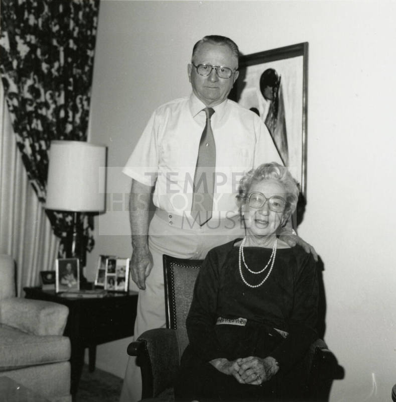 An unknown seated woman and standing man pose in front of a painting