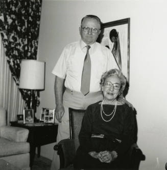 An unknown seated woman and standing man pose in front of a painting