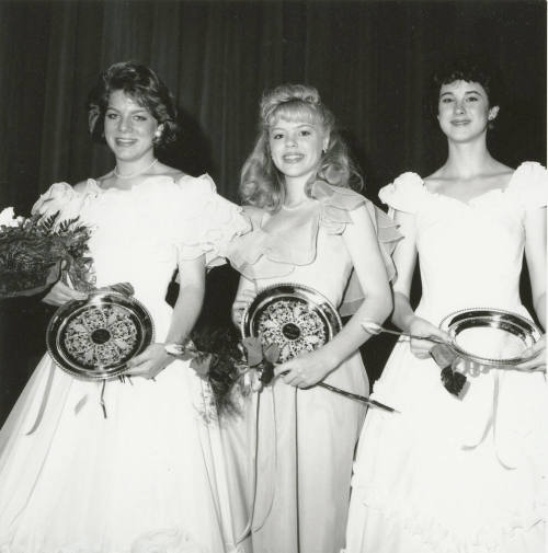 Three standing females wearing ballgowns each hold a bouquet and plate