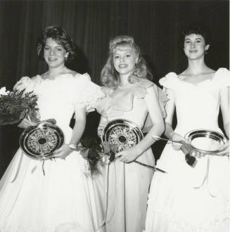 Three standing females wearing ballgowns each hold a bouquet and plate