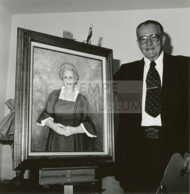 An man holding two paintbrushes stands next to a portrait of an unknown woman