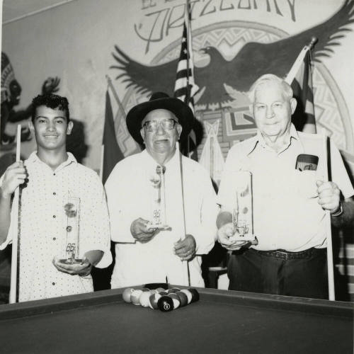Three unidentified men at a pool table with trophies