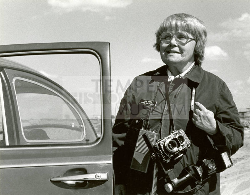 Jan Young with camera in her legendary raincoat beside her VW bug.