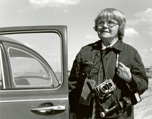 Jan Young with camera in her legendary raincoat beside her VW bug.