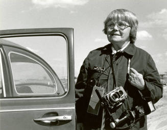 Jan Young with camera in her legendary raincoat beside her VW bug.