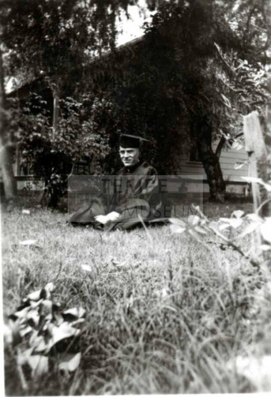 Frank Raymond in his cap and gown, sitting on a lawn in front of a house.