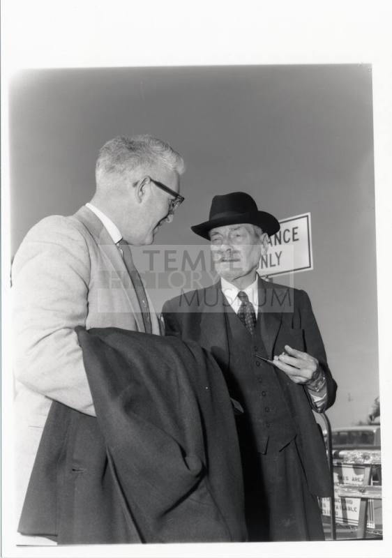 Clement Attley speaks with a man while smoking a pipe.