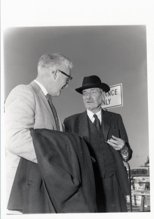 Clement Attley speaks with a man while smoking a pipe.