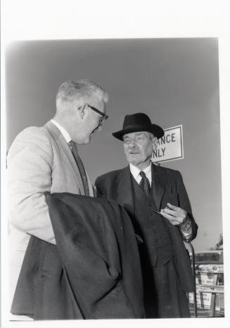 Clement Attley speaks with a man while smoking a pipe.