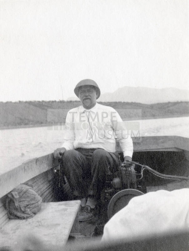 Snapshot of Dr. Fenn J Hart sitting in the back of a small wooden boat floating in an unknown body of water with mountains in the background