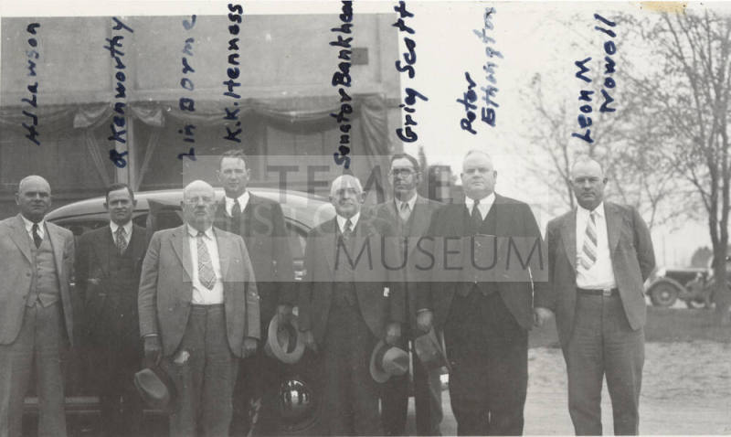 Photo- View of a identified group of eight men in front of a car and building