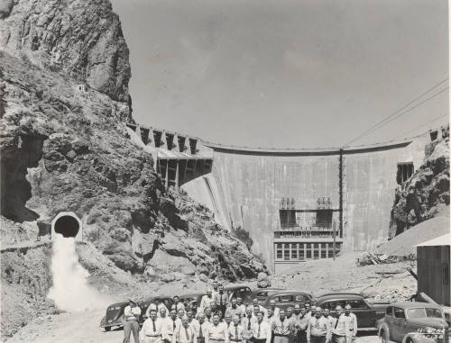 Photo- A row of thirty six men standing in front of Horse Mesa Dam with cars